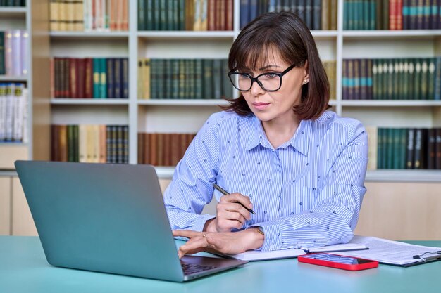 Middleaged female teacher working in library using laptop