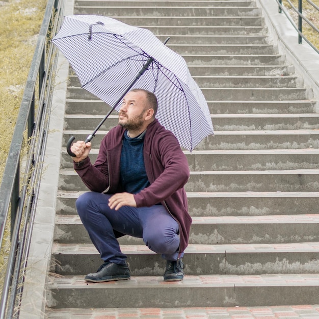 A middleaged man with a beard on the stairs under an umbrella