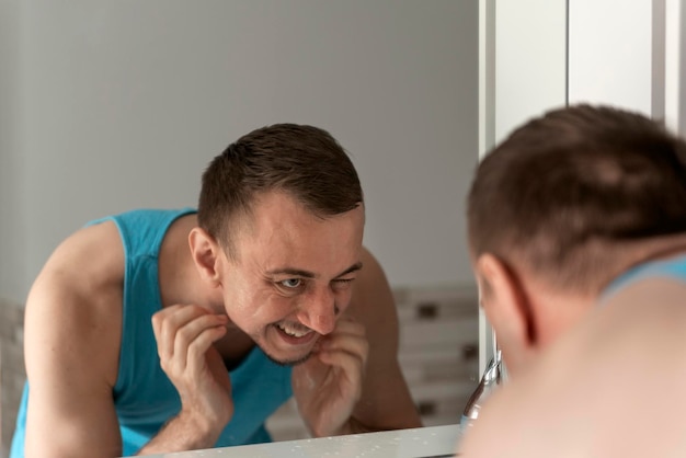 Middleaged man with no shaving face stands bent over sink unpleasure looks in mirror and scratch his bristle Morning hygiene