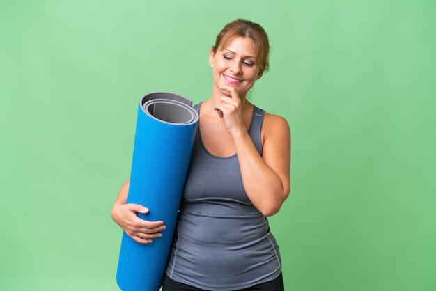 Middleaged sport woman going to yoga classes while holding a mat over isolated background looking to the side and smiling