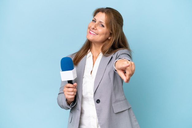 Middleaged TV presenter woman over isolated blue background pointing front with happy expression