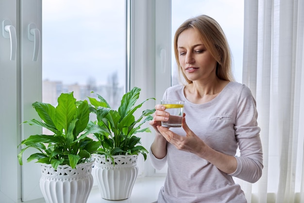 Middleaged woman drinking lemon water at home near the window