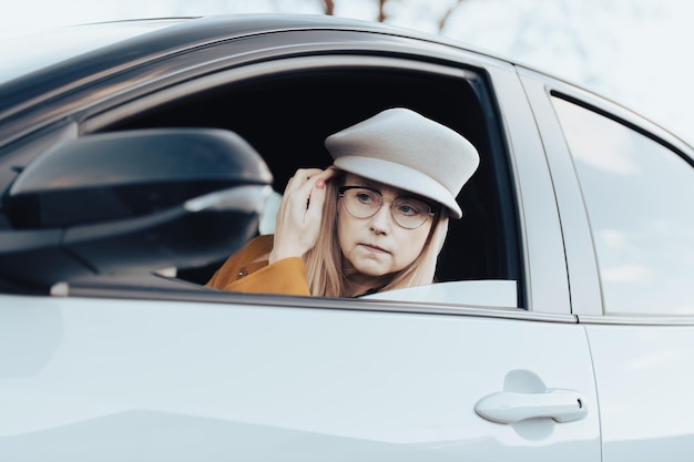 Photo a middleaged woman in glasses and hat sits in a car and watches her face in the rearview mirror