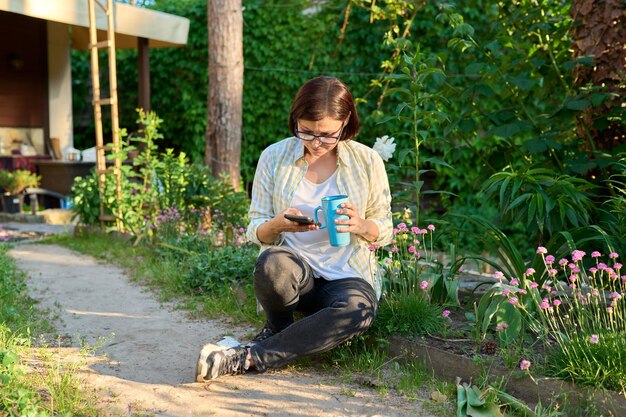 Middleaged woman having rest in spring garden with cup of tea and smartphone in hands