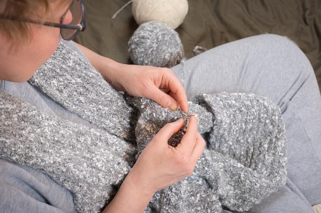 A middleaged woman is knitting a gray wool scarf The woman is doing needlework View from above