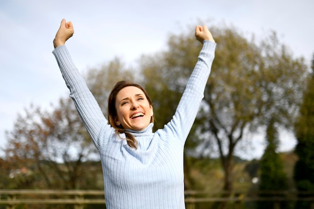 Middleaged woman in the park or field raising her arms in joy celebrating promotion or victory winning concept Autumn Winter roll neck jumper