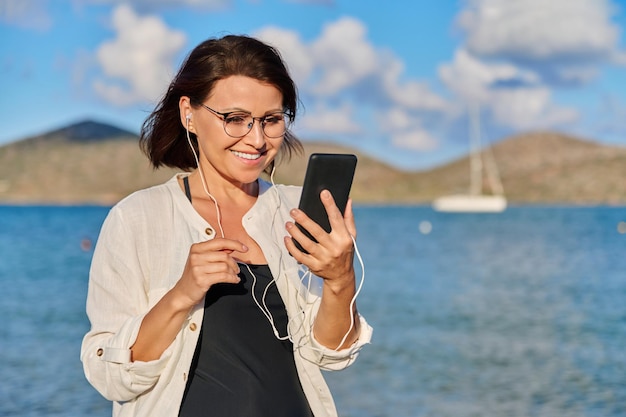 Middleaged woman relaxing on beach wearing headphones with smartphone