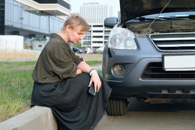 Middleaged woman sits on the side of road in front of brokendown car with an open hood