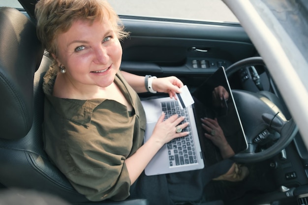 Middleaged woman sits behind the wheel of a car with a laptop on her lap