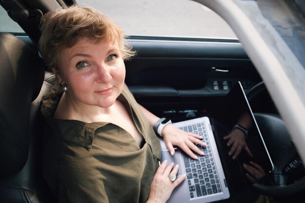 Middleaged woman sits behind the wheel of a car with a laptop on her lap