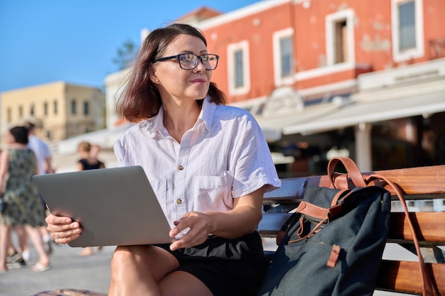 Middleaged woman sitting on a bench in city using a laptop