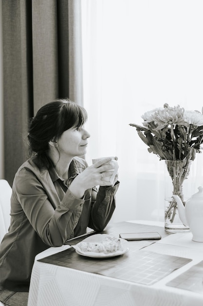 Middleaged woman sitting at the table in the cosy kitchen in the apartment and drinking tea with apple pie cozy autumn days black and white atmospheric photo