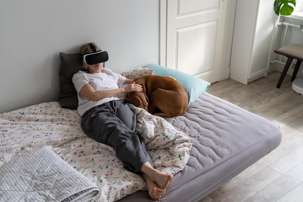 Middleaged woman spends free time playing VR games lying on bed near sleeping dog