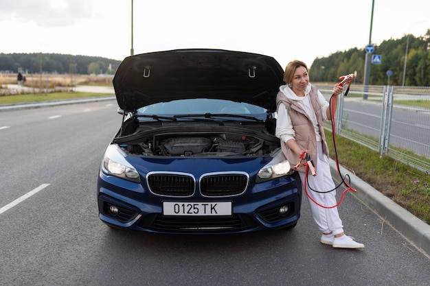 A middleaged woman stands at the hood of a broken car with wires for the battery and is waiting for