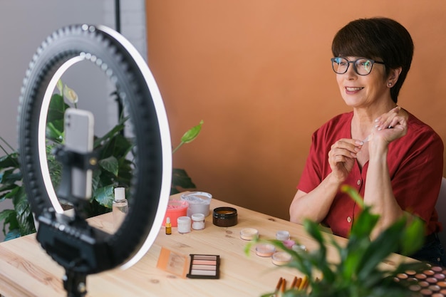 Middleaged woman talking on cosmetics with makeup eye shadows and blush palette and brushes while recording her video Mature female making video for her blog on cosmetics