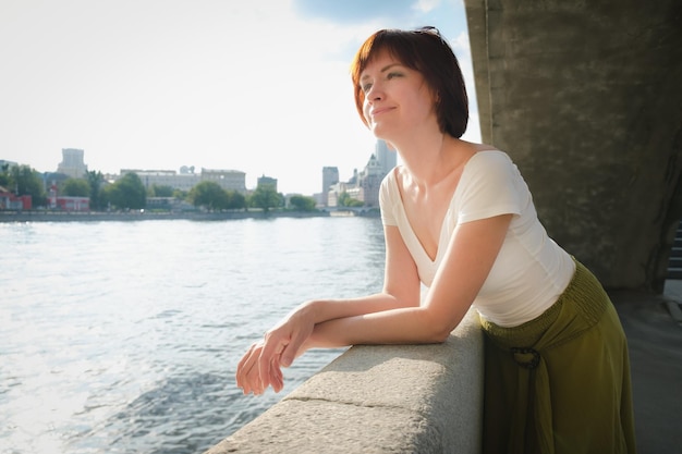 Middleaged woman with brown hair stands on embankment against the background of the urban landscape