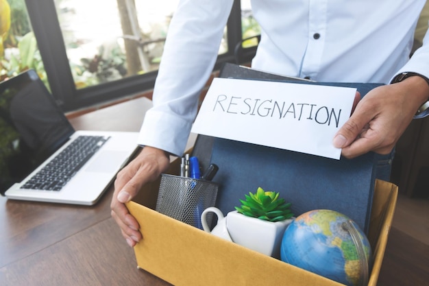 Photo midsection of businessman holding office supplies with resignation text in cardboard box