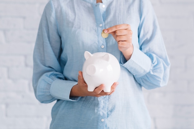 Midsection view of a woman's hand inserting coin in white piggybank