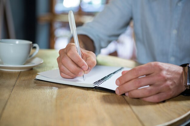 Midsection of young man writing in diary at table in coffee shop