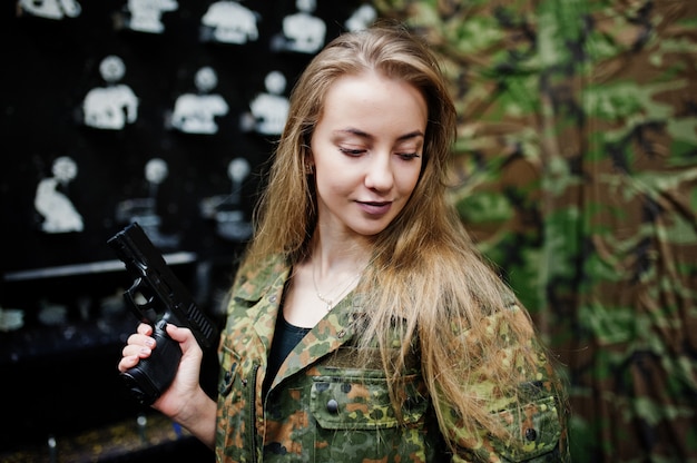 Military girl in camouflage uniform with gun at hand against army background on shooting range.