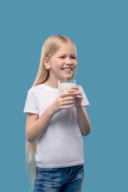 Milk for health. Smiling pretty girl with long blond hair standing holding glass of milk in her hands