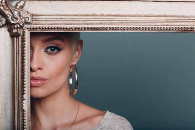 Millenial young woman with short blonde hair holds gilded picture frame in hands behind her face portrait.