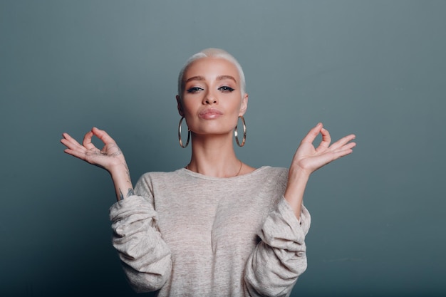 Millenial young woman with short blonde hair portrait sitting and meditate with ok sign mudra on gray blue background