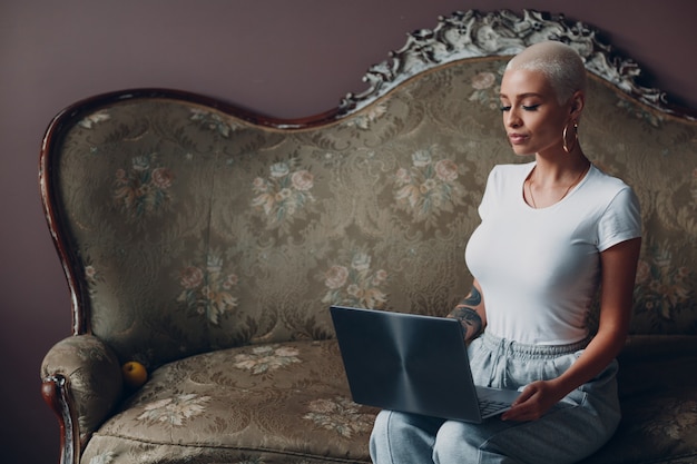 Millenial young woman with short blonde hair portrait sitting with laptop on vintage sofa at home of...