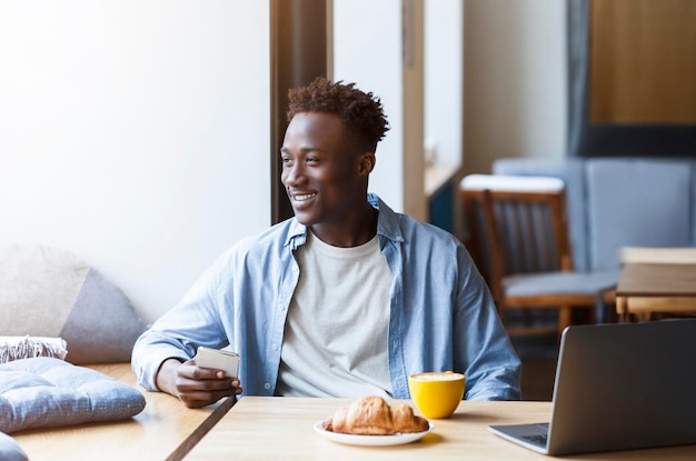 Millennial african guy with smartphone and laptop sitting in cafe