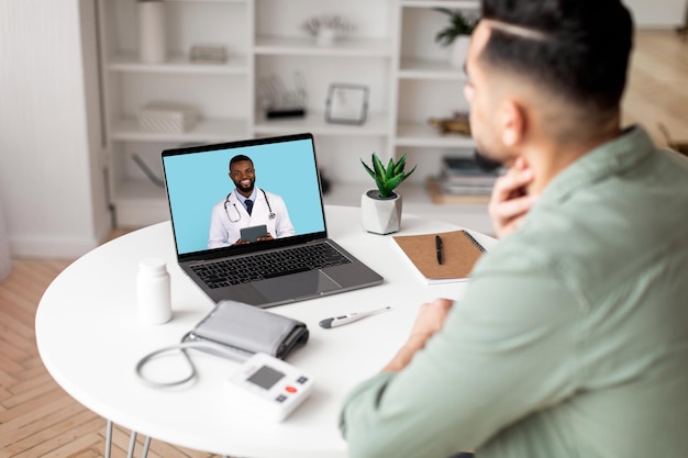 Millennial arab man checks throat calls online to doctor on laptop screen in living room interior