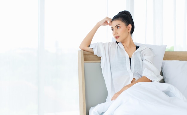 Millennial Asian sexy thoughtful female in casual comfort pajamas sitting leaning on pillow under white thick warm blanket thinking looking outside alone after waking up in bed in bedroom at home