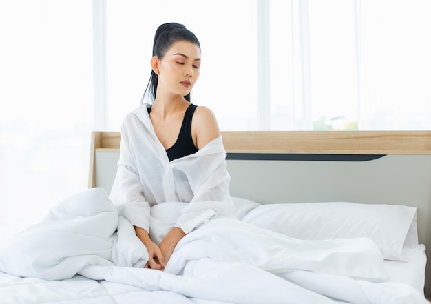 Millennial Asian sexy thoughtful female in casual comfort pajamas sitting under white thick warm blanket starting day thinking looking outside alone after waking up in bedroom in morning at home