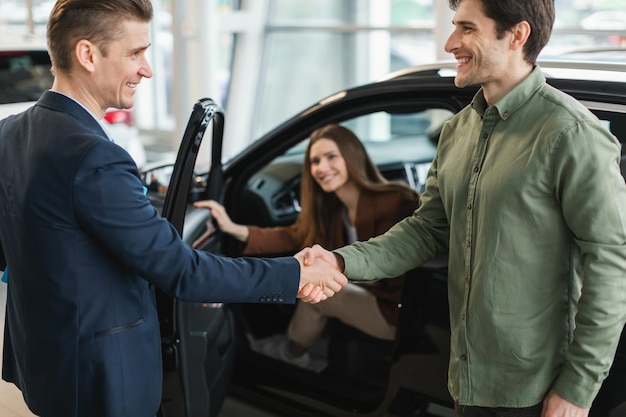 Millennial couple shaking hands with car salesman purchasing new vehicle at auto dealership