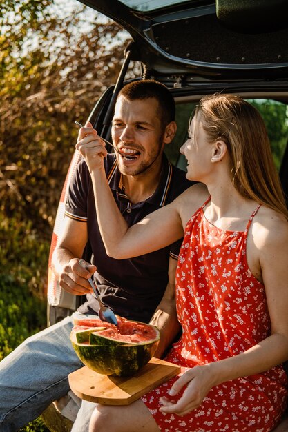 Millennial couple sitting on open trunk and eating watermelon Happy young couple having break at countryside