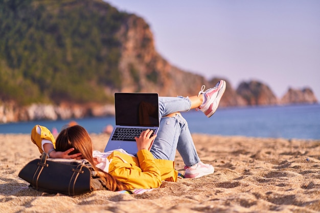 Millennial freelancer female using mockup laptop with empty screen blank and lying on the sand beach by the sea Dream office remote work concept