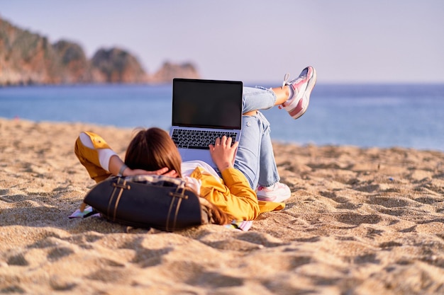 Millennial freelancer female using mockup laptop with empty screen blank and lying on the sand beach by the sea Dream office remote work concept