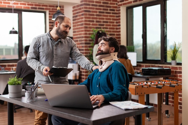 Millennial freelancer recovering after accident having neck pain wearing medical neck collar while working in brick wall startup office. Businessmen analyzing company turnover discussing business stra