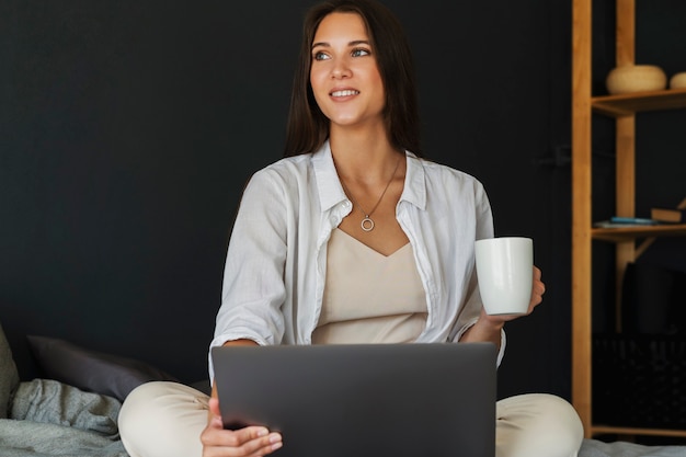 Millennial girl is sitting on bed in white shirt, holding cup of coffee in her hand.