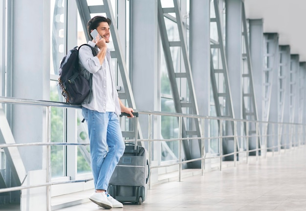 Millennial guy talking on phone waiting for flight with luggage at airport