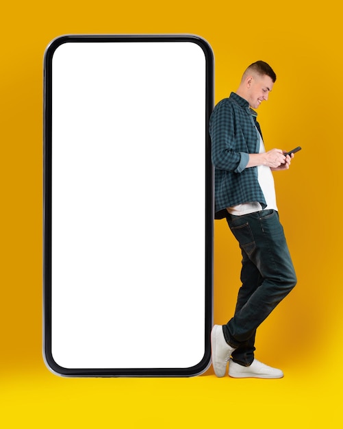 Millennial man using phone leaning on huge smartphone yellow background