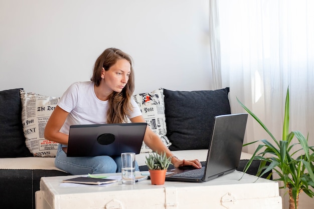 Millennial woman works at home,  seats close to Laptop computer with notebook, glass of water on white table in home workspace.