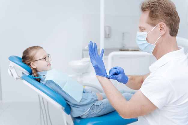 Mindful experienced dedicated dentist putting on medical gloves for protecting himself and his patient from any infection and following required instructions