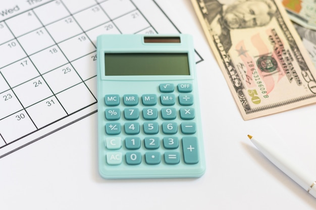 Mini calculator, blank calendar, pen and US dollars, 50 and 100. American cash, savings, taxes, benefit, income, money, finance. Top view, flat lay.