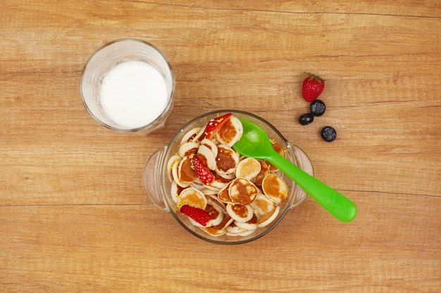 Mini pancakes with strawberries and blueberries in a glass bowl are on a wooden table next to a glass of milk