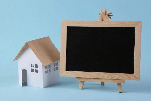 Mini paper house figurine and empty chalk board on a light blue background