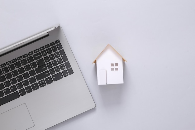 Mini paper house figurine and laptop on gray background Flat lay