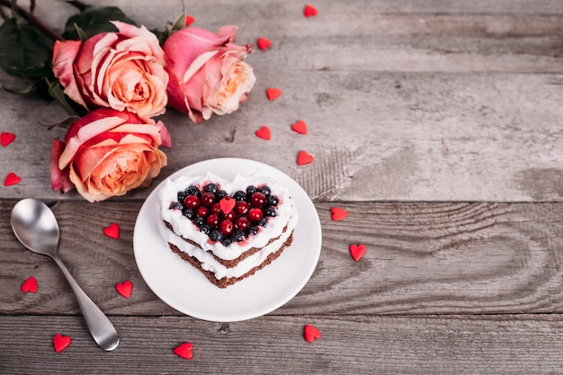 Mini romantic dessert cake for Valentine's Day with roses. Sweet cookies with cream topping and red heart for decor on wooden table. Close-up, copy space.