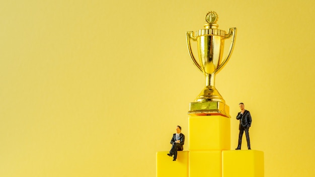 Miniature of businessman stands on podium ladder with golden trophy on yellow surface