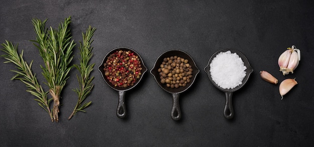 Miniature pans with spices salt black pepper and fragrant pepper a sprig of rosemary on a black table Spices for cooking