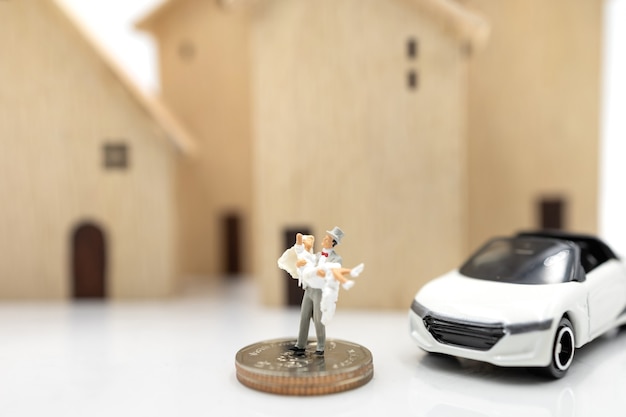 Miniature people: Couple in wedding dresses stand on coins stack with house and car. Family planning and housing concept.